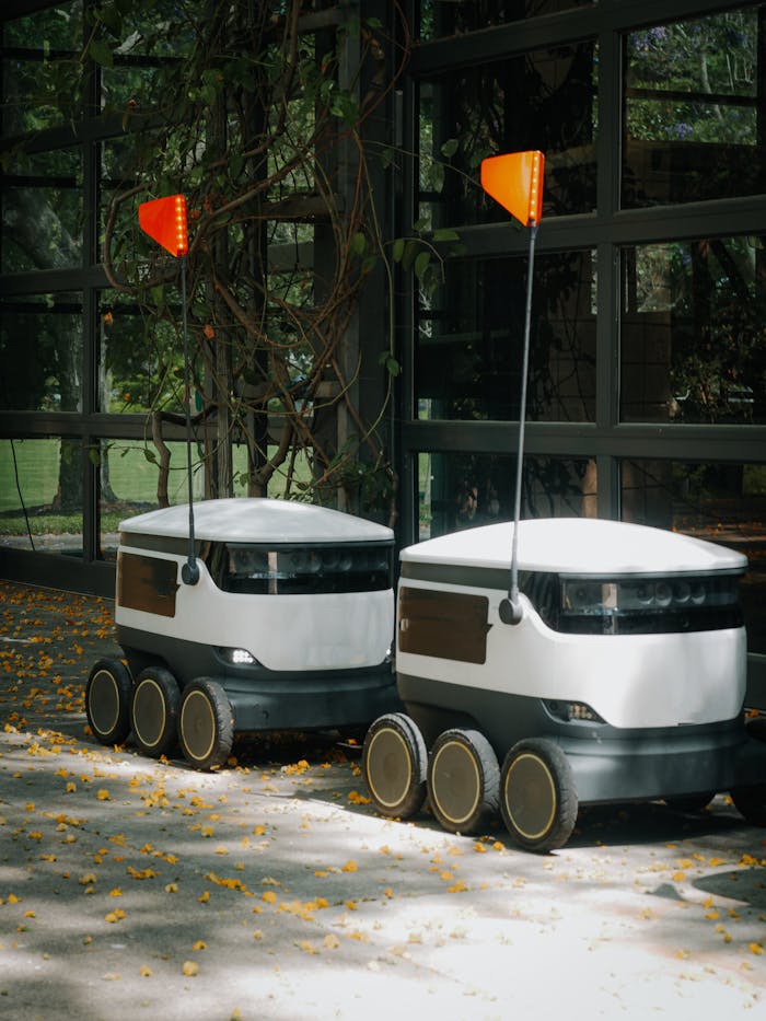Two modern delivery robots ready for autonomous operation in a green, leafy space.